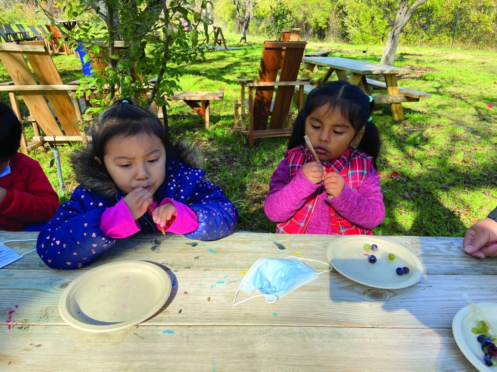 Two children sit at table in an outdoor setting with plates in front of them.