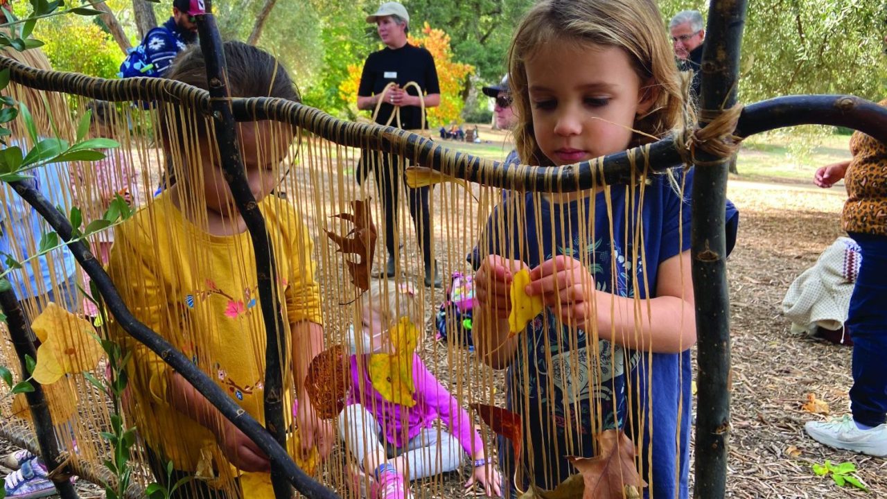 Two children stand in front of a loom like structure with threads hanging from wooden sticks and place leaves between the threads.