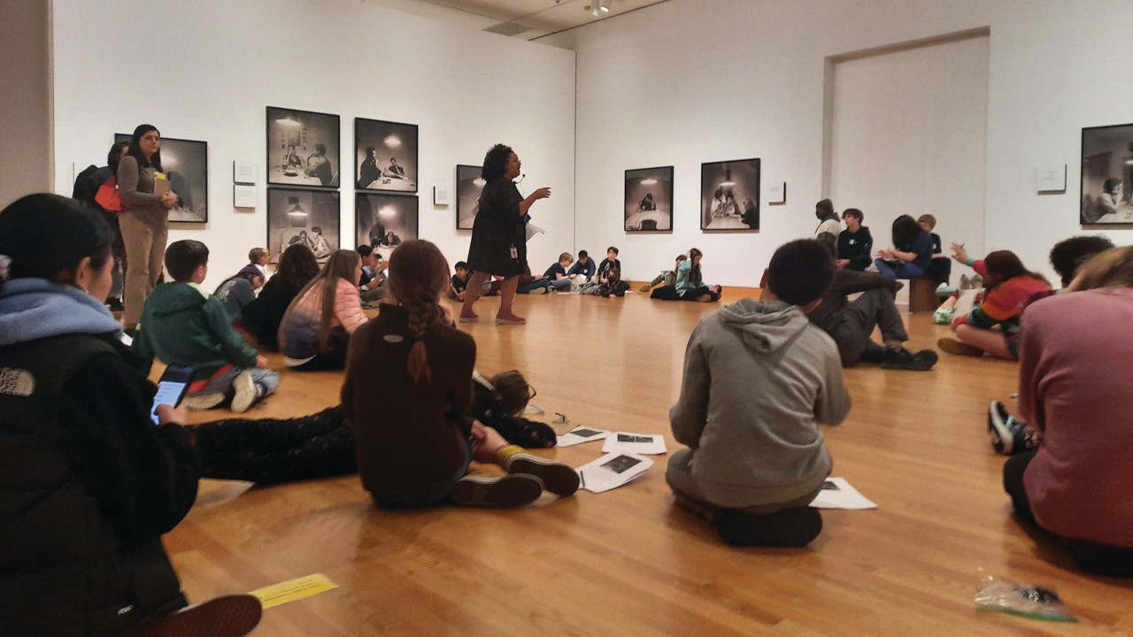 A group of people sit crossed legged in a circle on the floor in a museum gallery while an educator speaks.