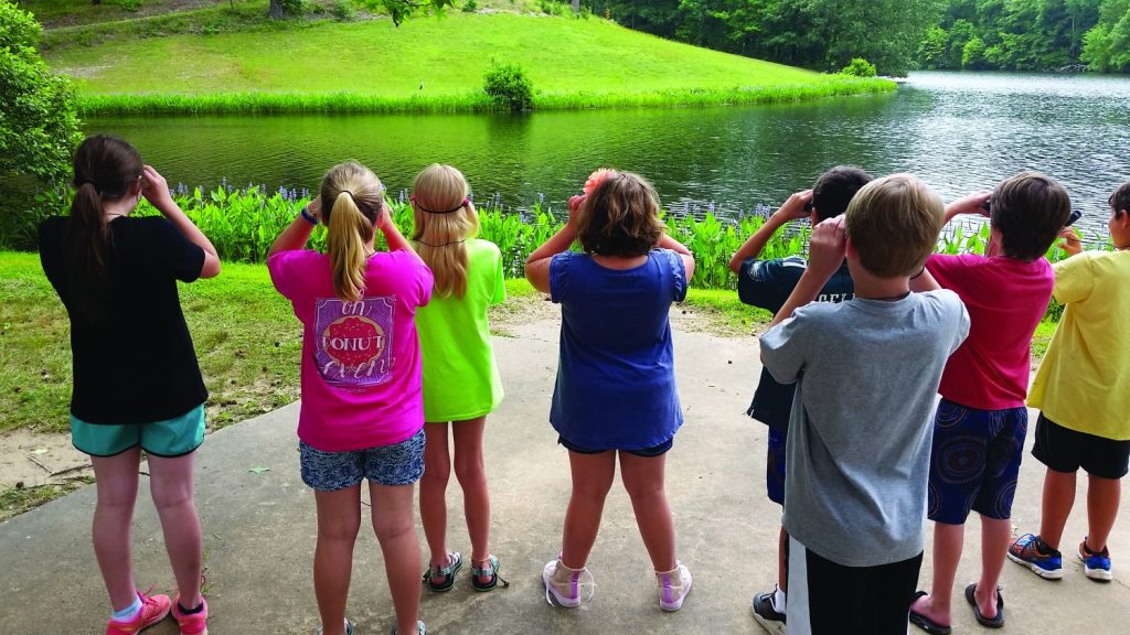 Several students stand next to a pond with their backs to the camera looking across the pond with binoculars. 