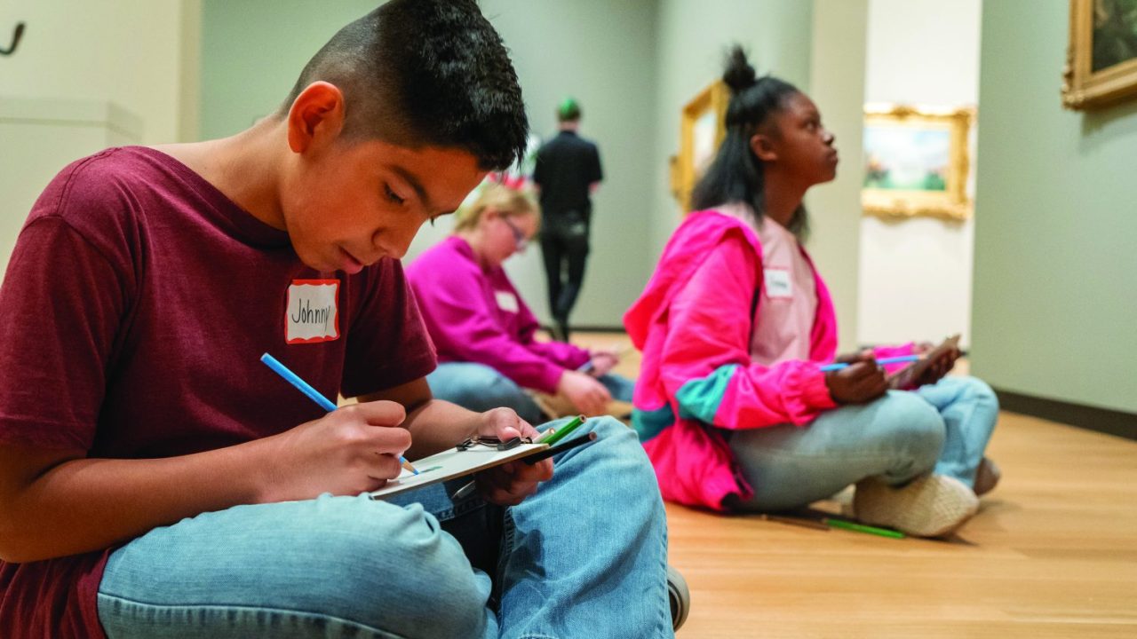 Three students sit crossed-legged on a wooden floor one is looking up at something above them, two others peer down at papers in front of them.