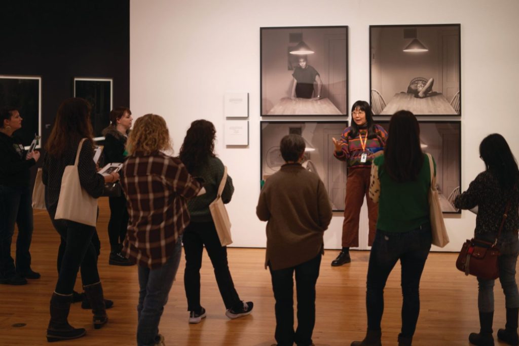 A group of people stand listening to an educator in front of a gallery with several framed images on the wall.