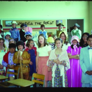 A group of children stand in various costumes behind small wooden desks in a classroom setting. 