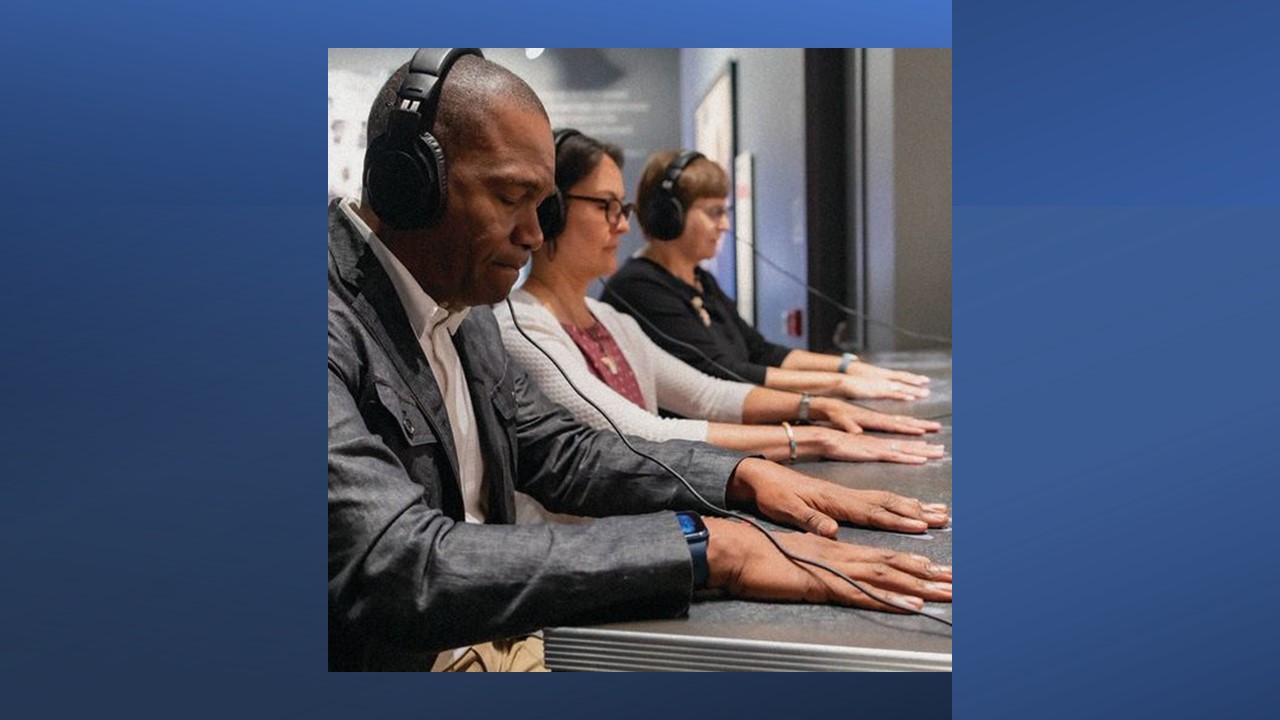 Three people sit at a table wearing headphones with their hands palm down on the table top.