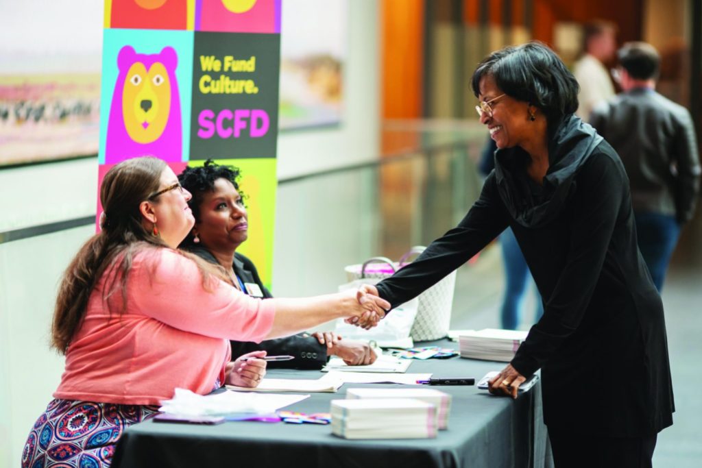 Two people sit at a table shaking hands with a third person standing across the table from them. 