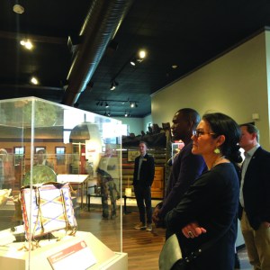 A group of people stand in a large gallery, one smaller group of two look at an object in an enclosed case in the foreground.