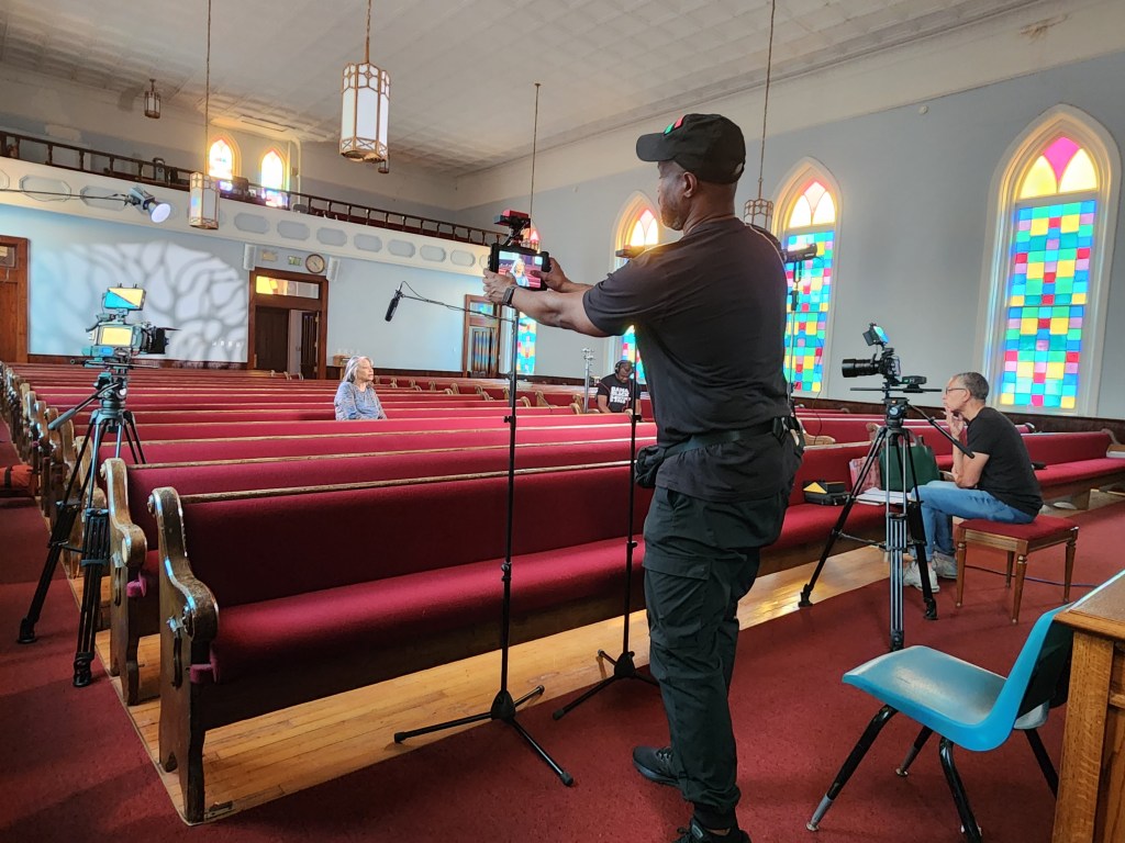 A film crew set up in a church with a subject seated in a center pew