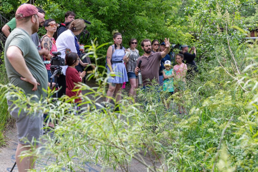 A group of people gathered around a riverbank, with one person speaking and gesturing.