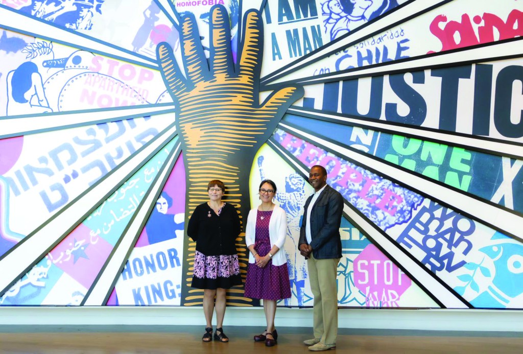 Three people stand facing the camera in front of a wall mural with a large outstretched hand.