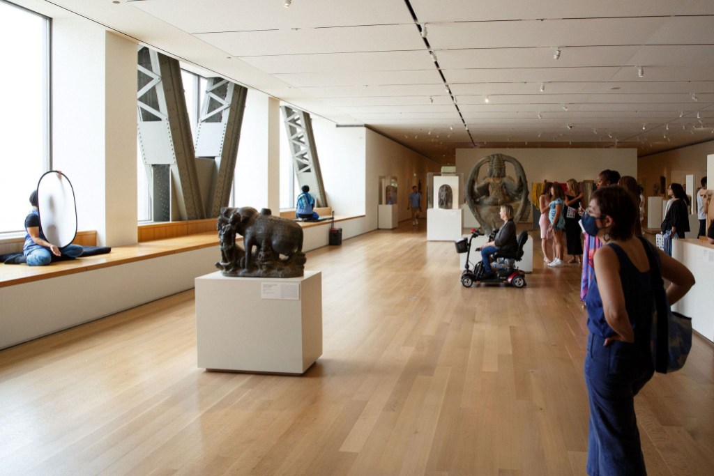 An audience watching performers seated on a bench in a museum gallery holding white screens in front of their faces