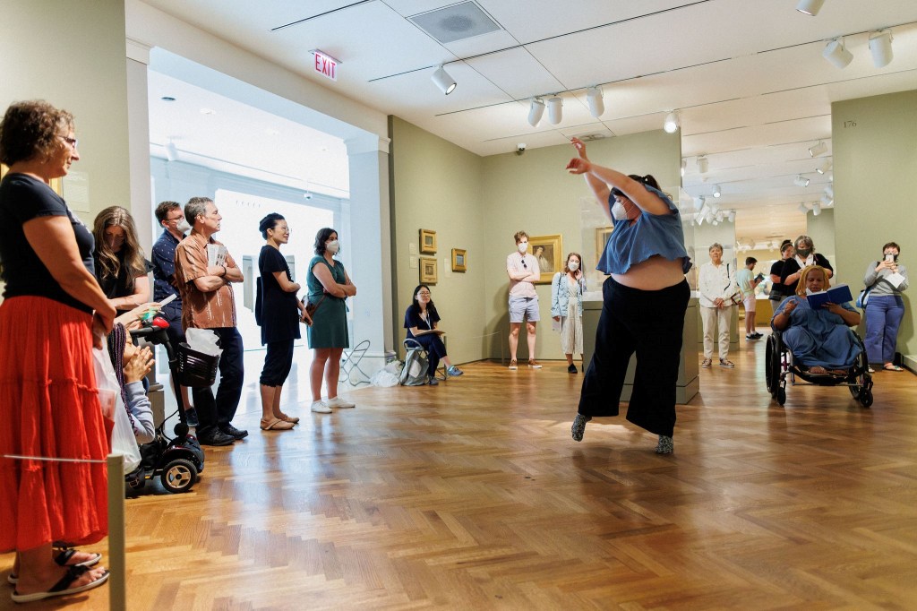 Visitors watching a performance in a museum gallery where one person reads from a book while seated in a wheelchair and another poses with their arms above their head