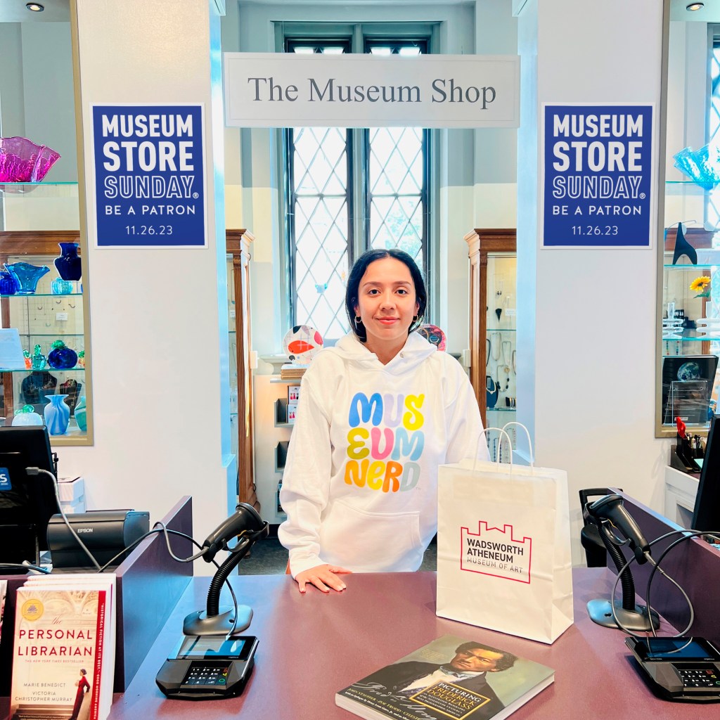 A museum shop attendant standing behind a counter while wearing a shirt that says Museum Nerd