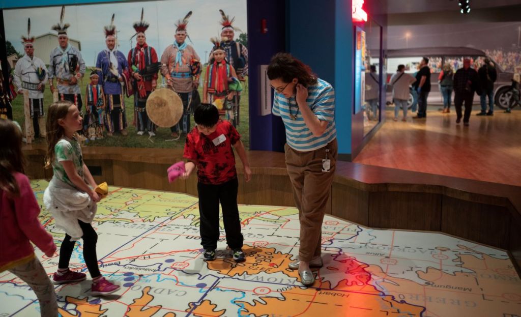 Two people stand on a map on the floor with a large image behind them of native American people.