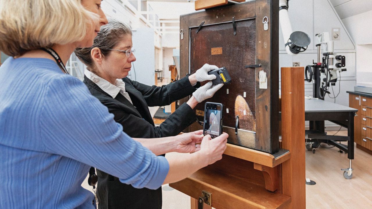 One person shines a light on the back of a framed piece while the other person takes a picture of the inscription.