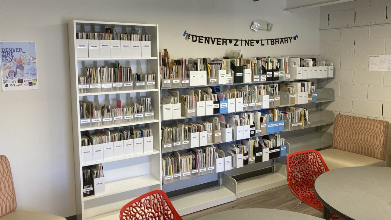 A room with a banner on the wall reading “Denver Zine Library,” with numerous tables and shelves stacked with books and other art supplies
