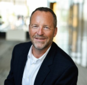 Middle-aged white male with dark hair, close-cropped mustache and beard, wearing a white shirt and dark blazer.