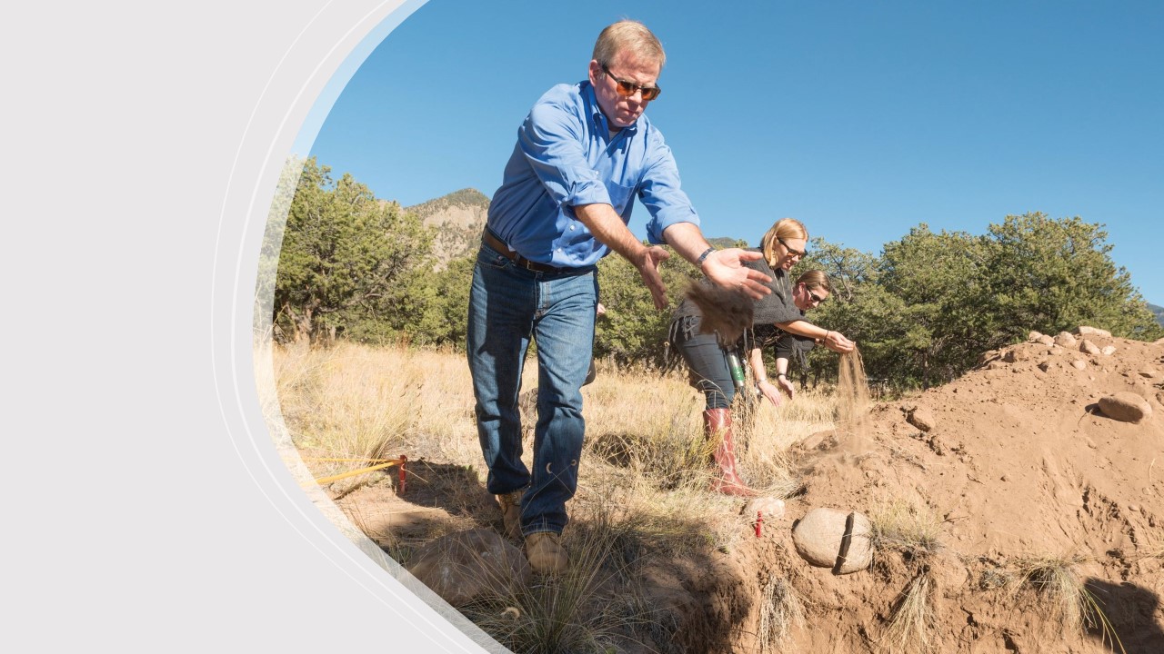 Three people stand on sandy ground spreading dirt over reinterred human remains.