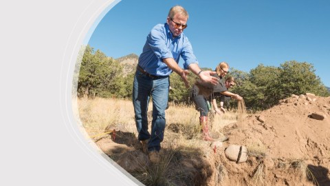 Three people stand on sandy ground spreading dirt over reinterred human remains.