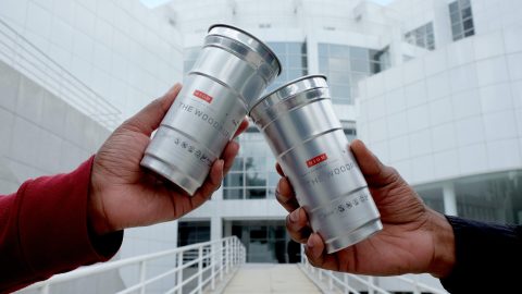 Two people holding up aluminum cups with the High Museum's logo on them in front of a building