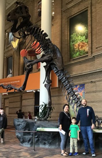 A three person family group stands in front of a dinosaur skeleton. 