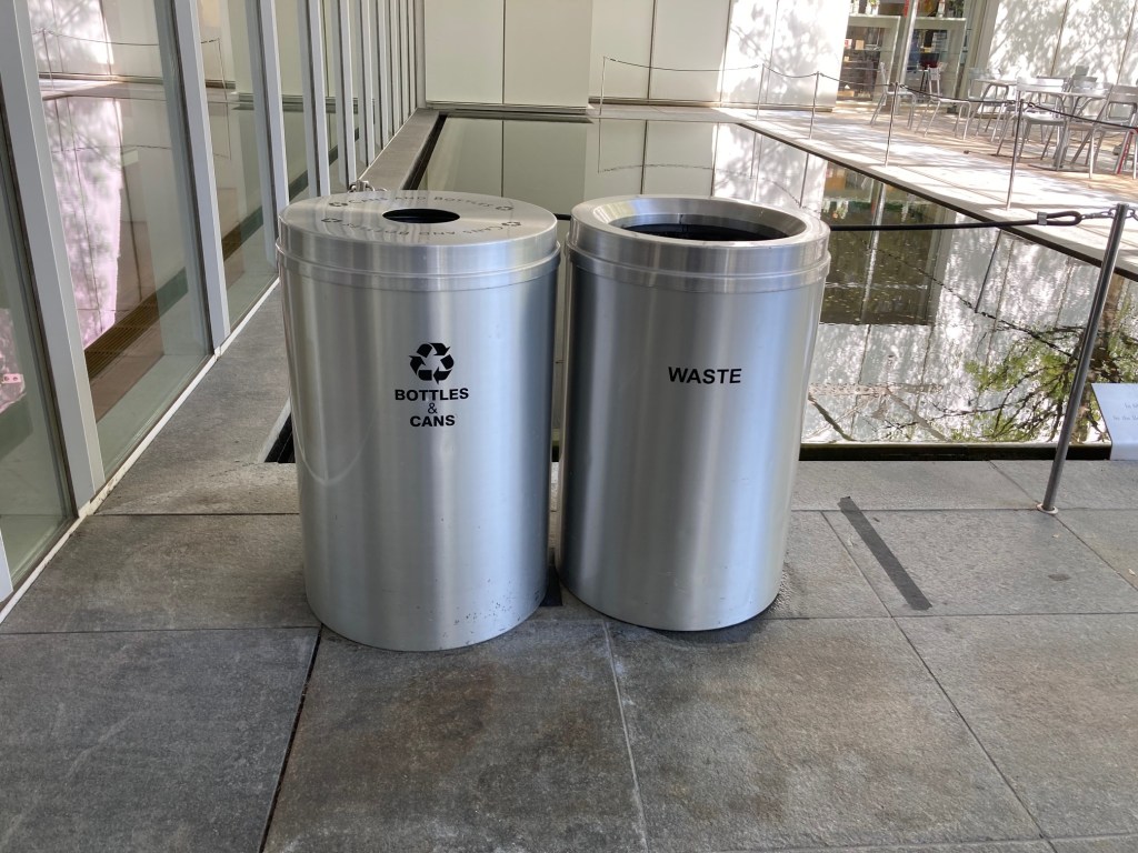 Two aluminum trash cans in a museum space, one reading "bottles and cans" with the recycling logo and the other reading "waste"