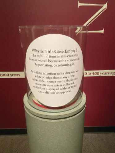 A round exhibit plinth stands with a round plexi vitrine that has a label reading, "Why is this case empty? The cultural item in this case has been removed because the museum is Repatriating, or returning it. By calling attention to its absence, we acknowledge that many of the cultural items once on display in our museum were taken, collected, studied, or displayed without Tribal consultation or approval."