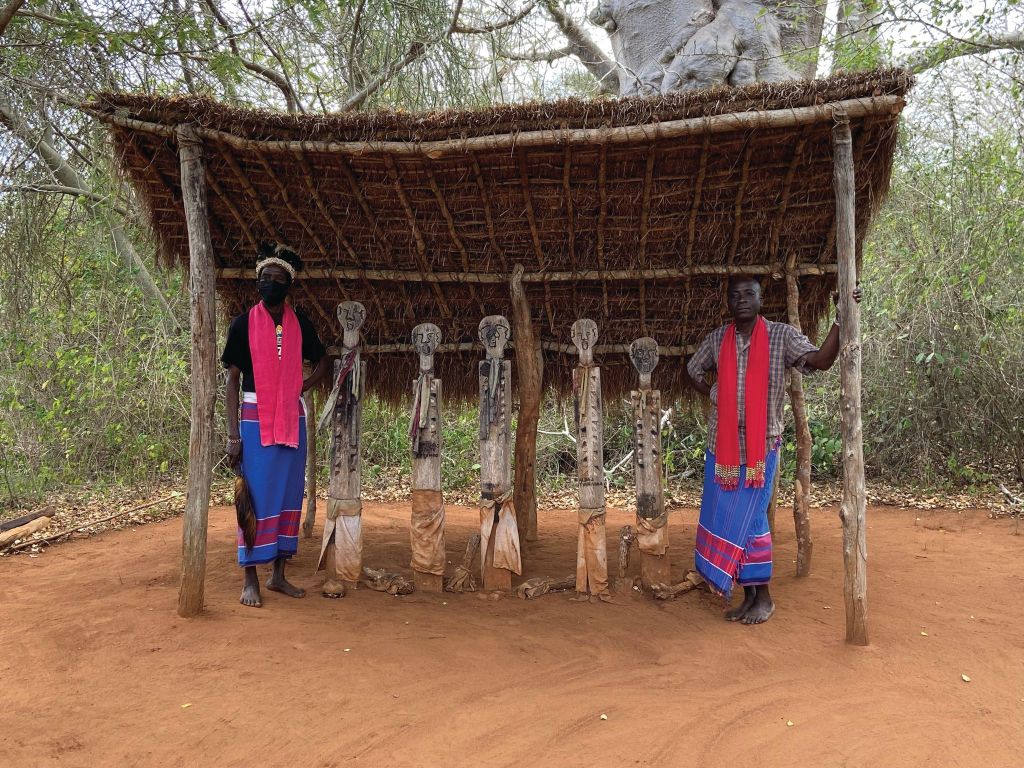 Two people stand under a lean to with a series of wooden statuary. 