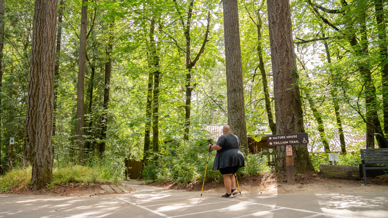 Back shot of a disabled Black non-binary hiker using trekking poles and walking from the parking lot onto the trail, framed by tall trees and forestry. They’re passing by a wooden sign with “Nature House” and “Trillium Trail ♿” marked ahead.