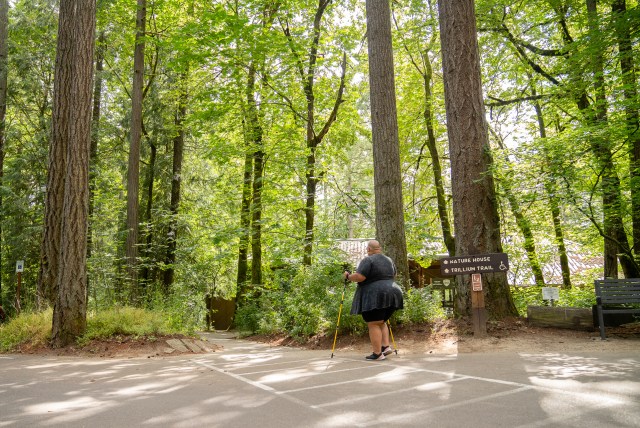 Back shot of a disabled Black non-binary hiker using trekking poles and walking from the parking lot onto the trail, framed by tall trees and forestry. They’re passing by a wooden sign with “Nature House” and “Trillium Trail ♿” marked ahead.