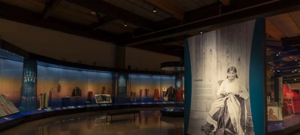 A museum gallery with textile objects behind glass and a wall panel with a photograph of a person in Indigenous clothing
