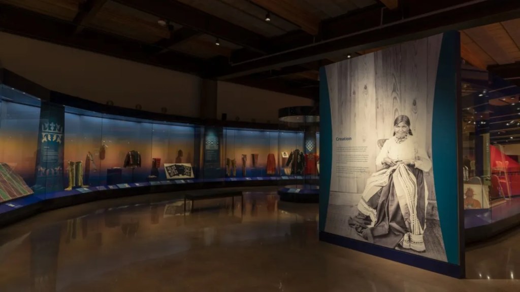 A museum gallery with textile objects behind glass and a wall panel with a photograph of a person in Indigenous clothing