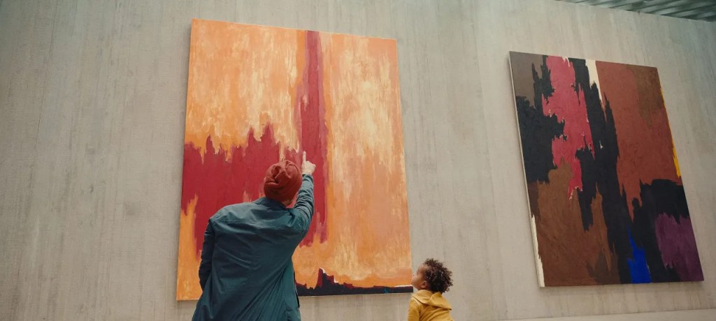 An adult and child kneeling on the floor of a museum and pointing at an abstract painting