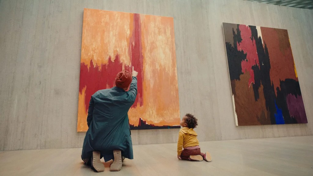 An adult and child kneeling on the floor of a museum and pointing at an abstract painting