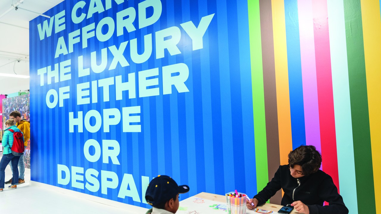 A person sits behind a desk in front of a colorful wall that has the words, "We cannot afford the luxury of either hope or despair."