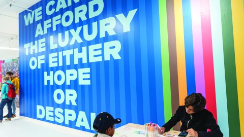 A person sits behind a desk in front of a colorful wall that has the words, "We cannot afford the luxury of either hope or despair."