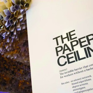 A giant paper flower with white and yellow petals that is attached to a ceiling and wraps around a wall that says, "The Paper Ceiling".