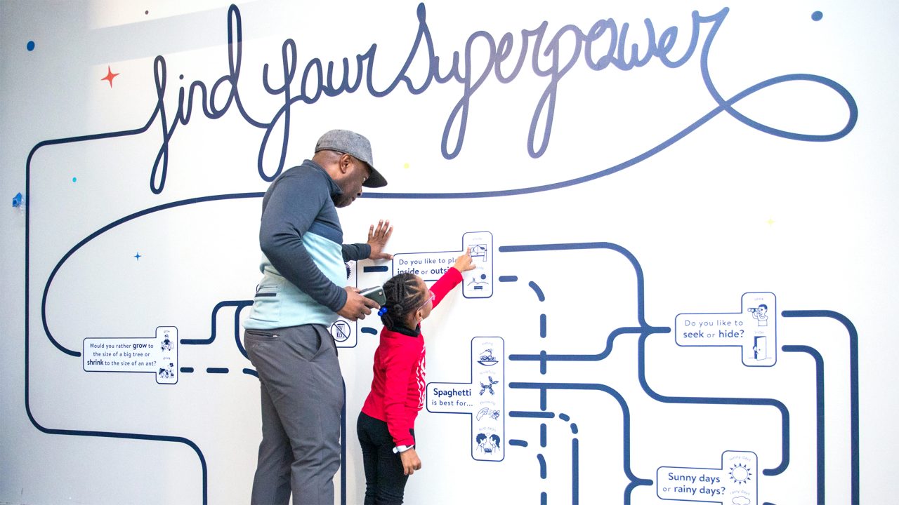 An adult and a child are standing together, studying a map painted on a wall