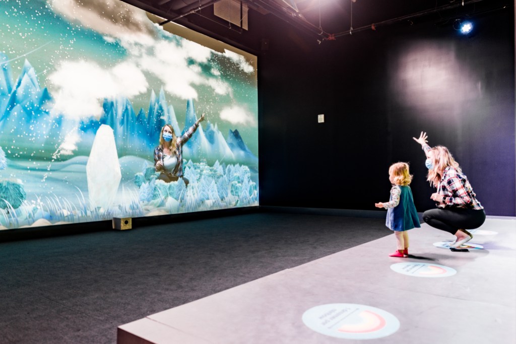 A child and adult standing in front of a large screen , engaged in an interactive activity