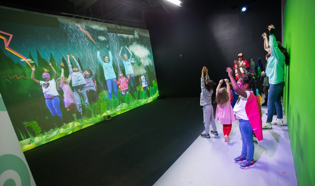A group of children with an adult chaperone posing in front of an augmented reality interactive that inserts weather imagery in front of them
