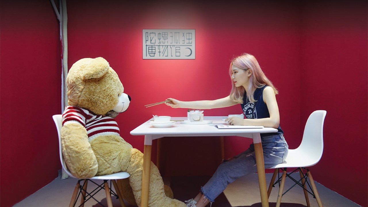 A young girl sits at a table in a red room across from a giant teddy bear.