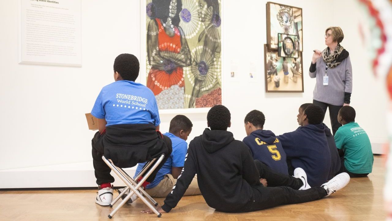 A group of children sitting in front of a painting as the teacher gestures towards the artwork.