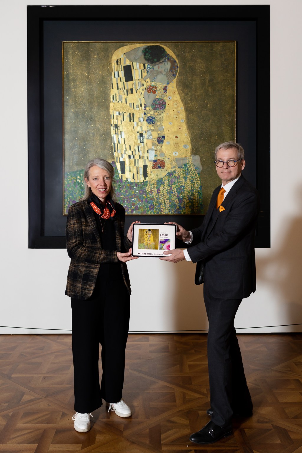 Two people posing in front of the physical "The Kiss" painting holding up a tablet showing the NFT