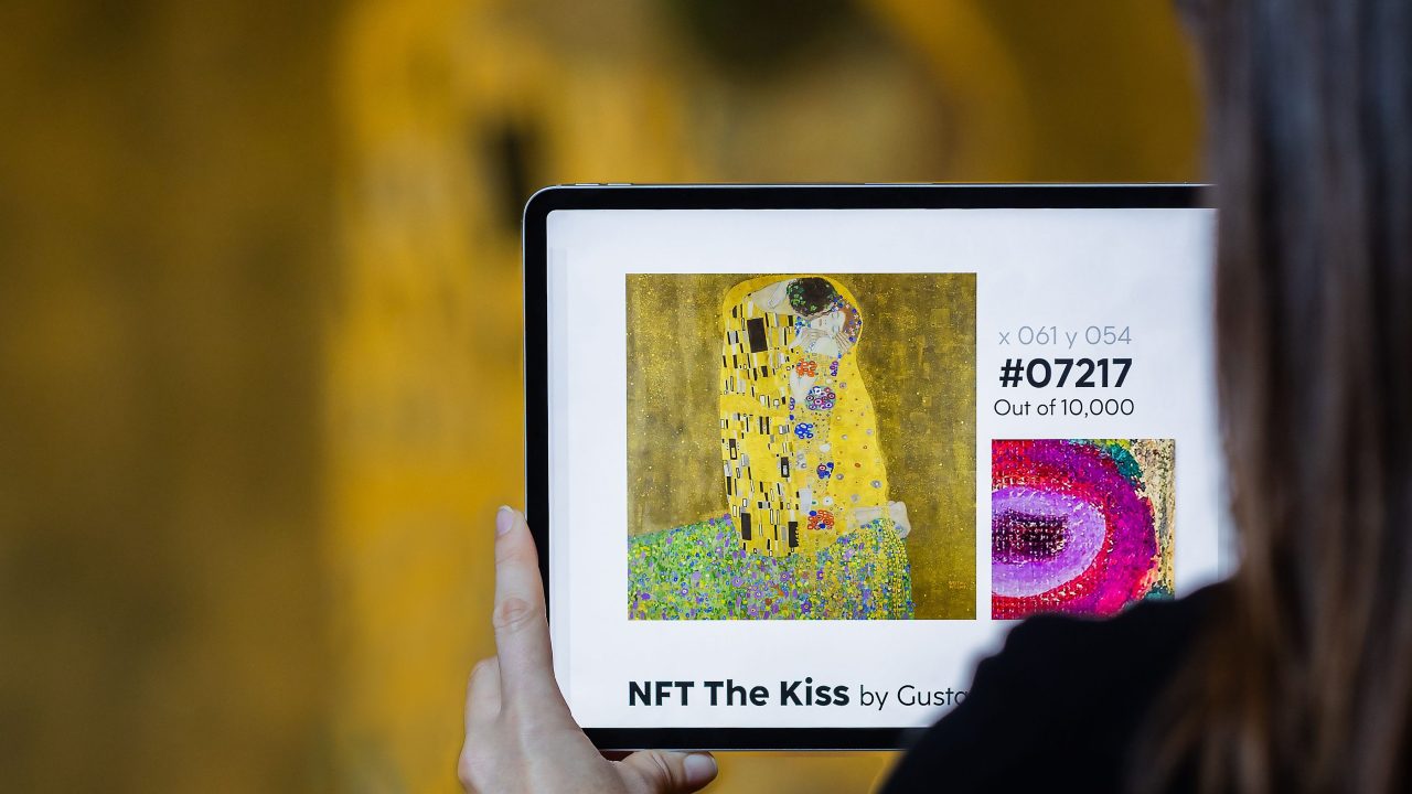 A person holding a tablet showing an image of Gustav Klimt's painting "The Kiss," with the text "NFT The Kiss by Gustav Klimt" and a blown-up title of the painting labeled "x 061 y 054 #07217 Out of 10,000"