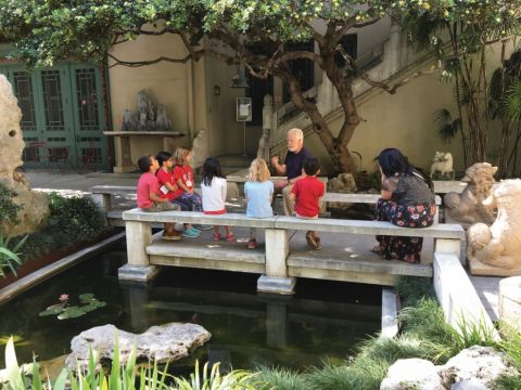 A guide sits outside in a garden setting with a group of children scattered around listening.