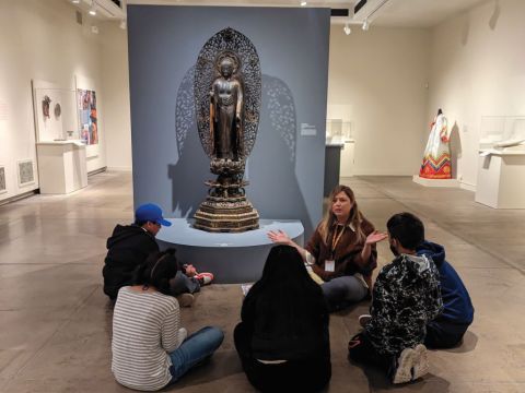 A gallery guide sits on the floor in an exhibition with a group of children scattered around listening.