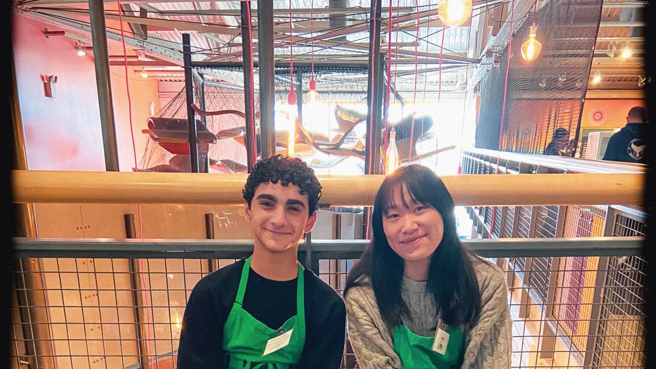 Two young people sit at a table with a green tablecloth both wearing green aprons ready to welcome people to their table of interactive exhibition elements.