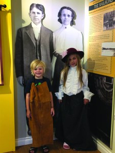 Two young children stand in front of a cardboard cutout wearing period clothing.