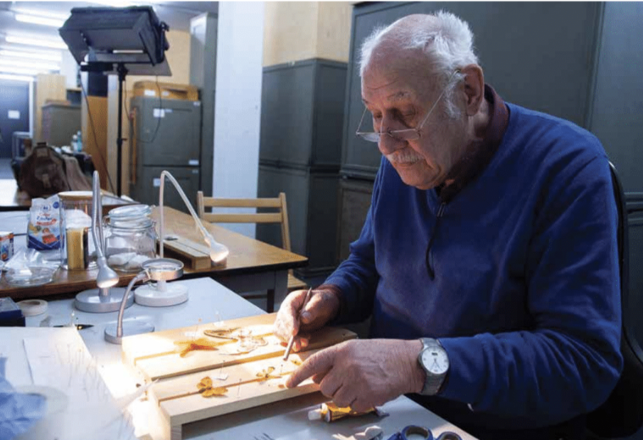 An elderly man stis at a table wearing reading glasses working on an object 