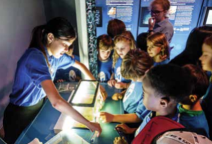 A group of students stand at a terrarium set up on a table where a volunteer describes the animal inside. 