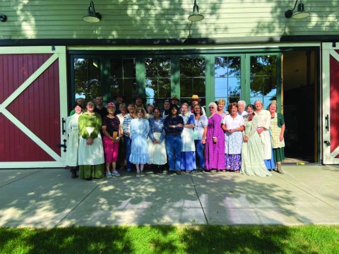 A large group of volunteers stand outside the barn wearing period dress.